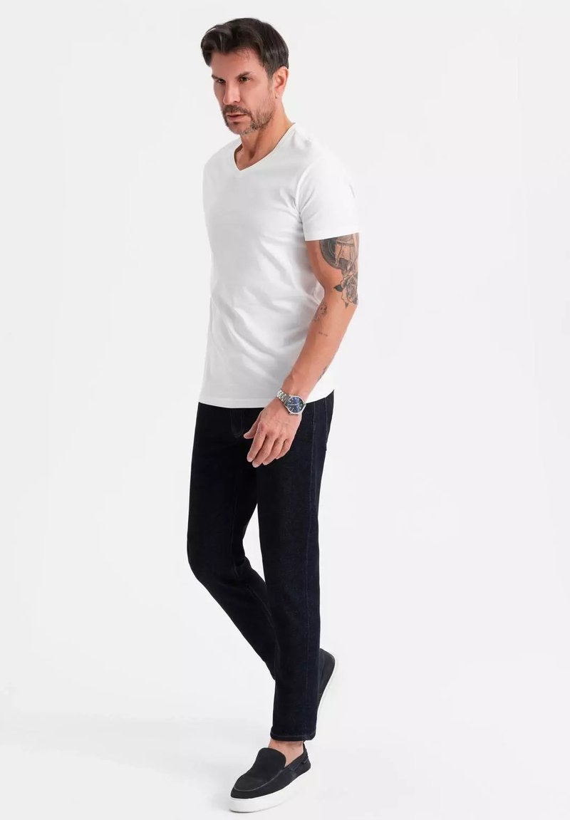 T-shirt bianco a manica corta con scollatura a V, jeans in denim scuri, scarpe slip-on nere. Presenta una vestibilità maschile e un tatuaggio sull'avambraccio, posato su uno sfondo neutro.