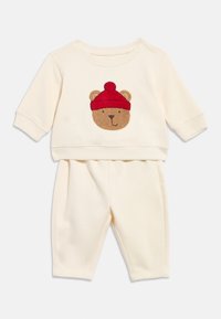 Cremefarbenes Baby-Outfit mit langen Ärmeln und Hosen, mit einem Teddybärgesicht, das eine rote Mütze trägt, auf dem Oberteil.