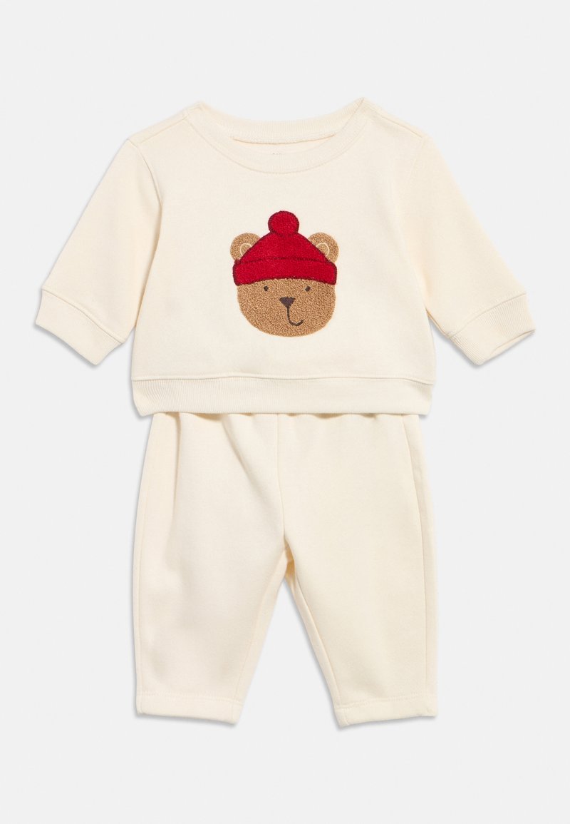 Cremefarbenes Baby-Outfit mit langen Ärmeln und Hosen, mit einem Teddybärgesicht, das eine rote Mütze trägt, auf dem Oberteil.