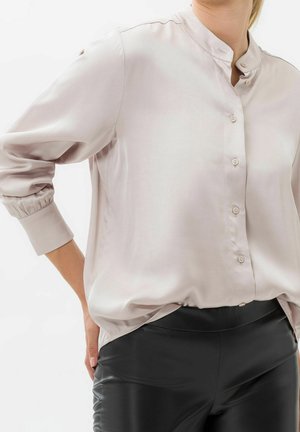 Frau trägt hellbeige Bluse mit Stehkragen und schwarzen, hoch taillierten Hosen, Hand auf der Hüfte ruhend, vor weißem Hintergrund.