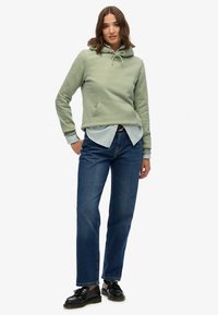 Sudadera verde claro con cordón, bolsillo frontal tipo canguro, llevada sobre una camiseta de rayas azules y blancas, combinada con jeans de mezclilla oscura y zapatos negros.