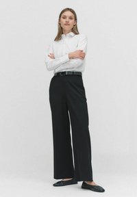 Witte blouse met lange mouwen, gecombineerd met hoge, zwarte wijde broek en zwarte platte schoenen. Strak, modern ontwerp.