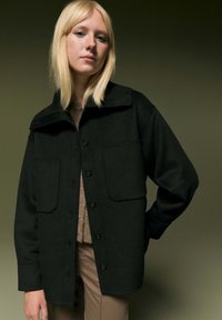 Veste en laine noire avec un col, des boutons à l'avant et deux grandes poches plaquées. Le modèle porte un haut beige en dessous. Texture douce visible.