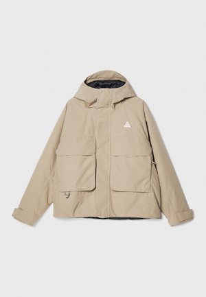Beige wasserdichte Jacke mit Kapuze, zwei großen Fronttaschen, verstellbaren Manschetten und einem Logo. Glattes Material mit robustem Design.