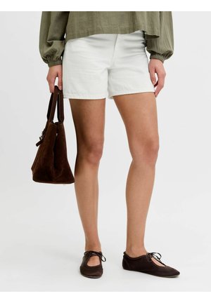 Žena nosiaca biele šortky, olivovo zelenú blúzku, tmavohnedé šnurovacie baleríny, drží tmavohnedú semišovú kabelku pri sebe.