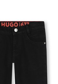 Jeans en denim noir avec bouton et rivets argentés, ceinture intérieure rouge arborant le texte "HUGO | 677", poche avant visible sur le côté droit.