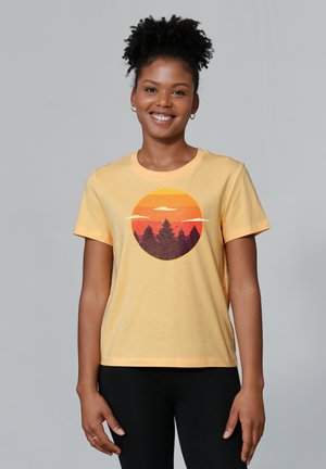 T-shirt en coton jaune avec un graphique de coucher de soleil circulaire aux tons orange et violet, contrasté par une silhouette d'arbres à feuilles persistantes.