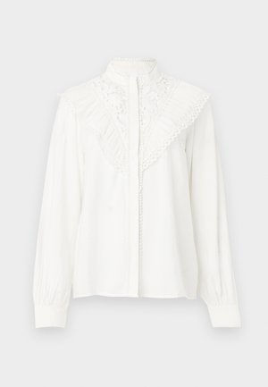Hvit langermet bluse med høy krage, med spiss detaljer, volanger på skuldrene og knappelukking foran. Kvalitets stofftekstur.