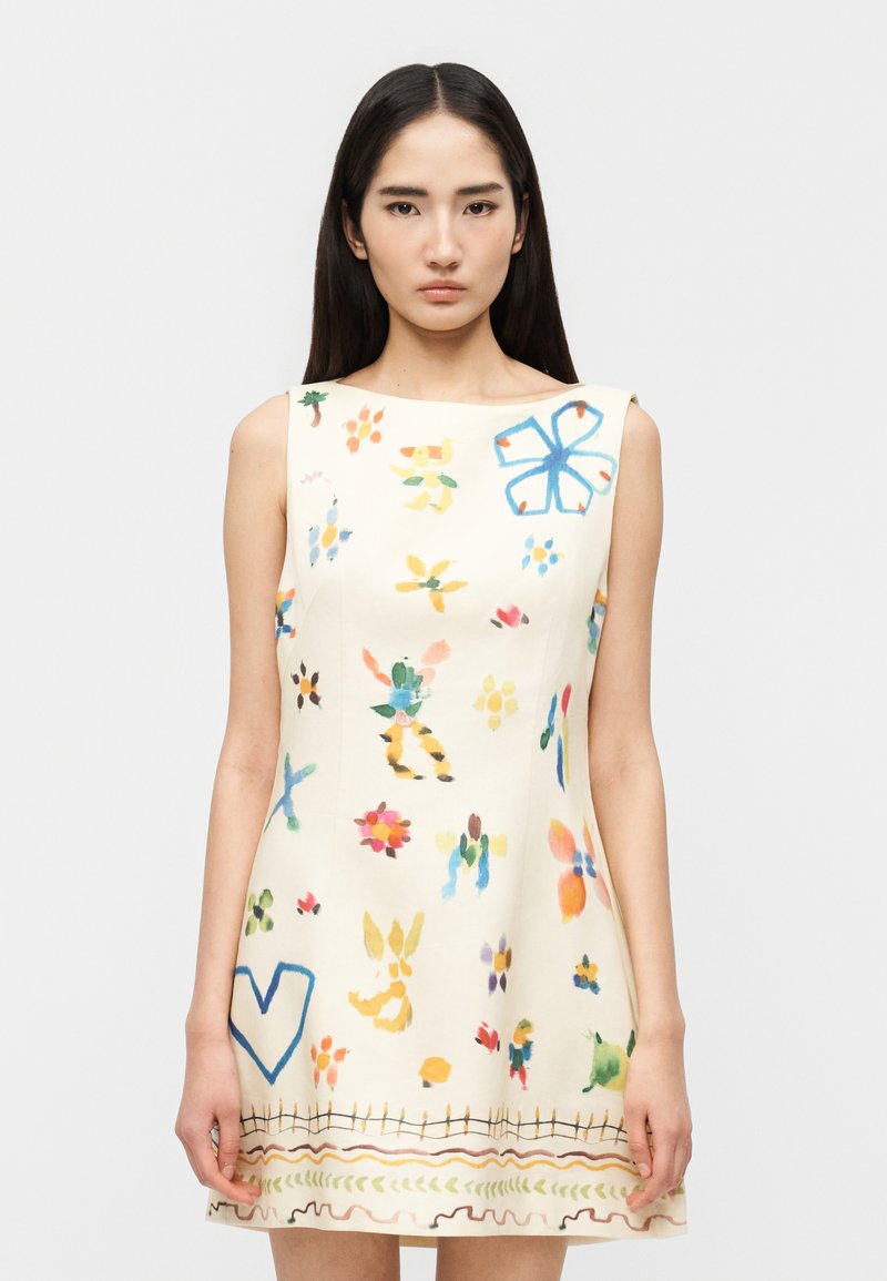 Νεαρή γυναίκα με μακριά σκούρα μαλλιά φοράει αμάνικο κρεμ φόρεμα με πολύχρωμα αφηρημένα μοτίβα λουλουδιών και καρδιών.