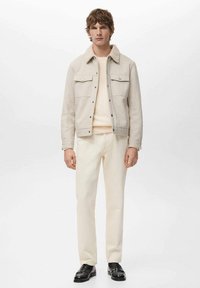 Hellbeige Jacke aus strukturiertem Stoff, mit Fronttaschen und Kragen; kombiniert mit einem leichten Pullover und cremefarbenen Hosen. Schwarze Schuhe vervollständigen das Outfit.