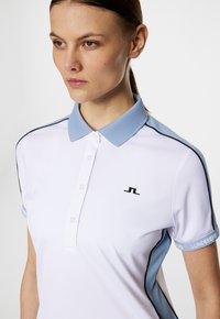 Vit polo tröja med ljusblå krage och detaljer, med svart logotyp på bröstet, korta ärmar med accentdesign och knappstängning.