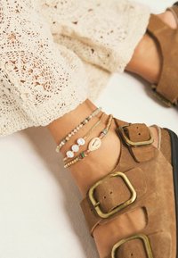 Sandali alla caviglia in pelle scamosciata marrone con fibbie dorate, indossati con bracciali sovrapposti contenenti perle e perline colorate su uno sfondo di pizzo.