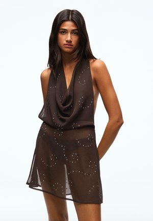 Ung kvinde med langt mørkt hår iført en gennemsigtig sort kjole med draperet halsudskæring og spredte små glitrende pynt.