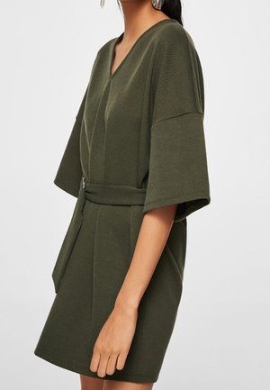 Femme portant une robe courte à manches courtes de couleur vert olive avec ceinture et col en V, debout devant un fond clair uni.