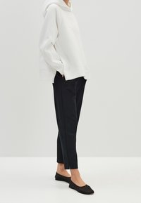 Personne portant un sweat à capuche blanc oversized, un pantalon noir fuselé à la cheville et des ballerines noires, debout les mains dans les poches.