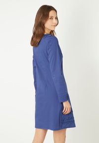 Vestido azul de manga larga hecho de una tela suave. Presenta un cuello, mangas texturizadas y un parche decorativo con lazos en el lado izquierdo.