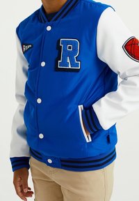 Modrá varsity bunda s bílými rukávy, zdobená filcovou nášivkou 'R' a emblematem basketbalu. Hlavice a pásky jsou žebrované, zapínání na knoflíky.
