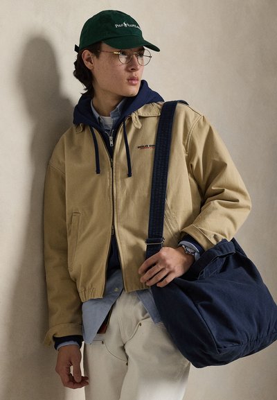 Νεαρός άνδρας φορώντας μπεζ μπουφάν, μπλε σκουρόχρωμη ζακέτα με κουκούλα, πράσινο καπέλο, γυαλιά, λευκό παντελόνι και κρατώντας μια μεγάλη μπλε τσάντα ώμου.