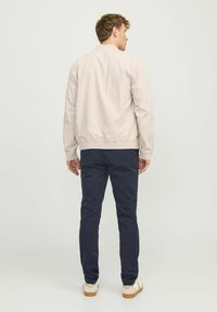 Helles beiges Bomberjacke mit gerippten Bündchen, getragen über dunkelblauen Chinos. Das Outfit besteht aus lässigen Schuhen mit leichter Sohle und einer strukturierten Oberfläche.