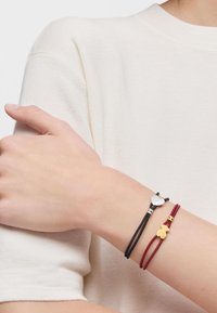 Dos pulseras con un corazón plateado y un charm de oso amarillo, hechas de cordones elásticos en negro y burdeos, llevadas en una muñeca de piel clara.