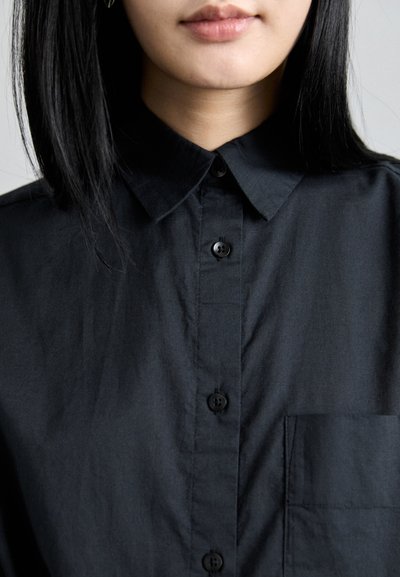 Black button-up shirt featuring a classic collar, front pocket, and smooth texture. Dark buttons contrast with the fabric.