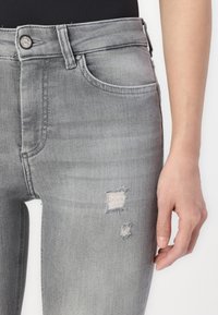Jeans de mezclilla gris con un acabado desgastado, que presentan detalles rasgados y parches deshilachados, cinco bolsillos y un diseño de cierre de botón.