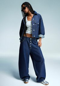 Denim jakna in široke hlače v temno modri barvi, z srebrnimi gumbi in zavihanimi manšetami, nosi čez belo majico brez rokavov. Srebrni čevlji dopolnjujejo videz.