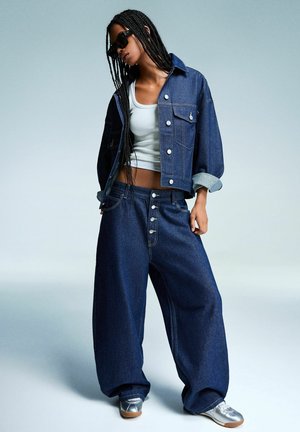 Denim jacket and wide-leg jeans in dark blue, featuring silver buttons and rolled cuffs, worn over a white tank top. Silver shoes complete the look.