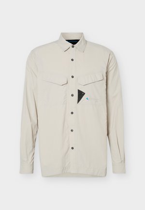 Chemise beige à manches longues avec boutons, dotée de deux poches poitrine, dont une avec un petit détail triangulaire noir et un logo subtil.