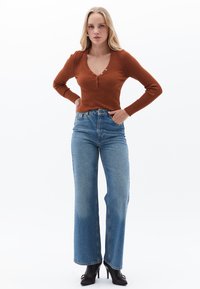 Top de manga larga acanalado en color óxido con escote en V y detalle de botones, combinado con jeans de tiro alto de mezclilla azul clara y zapatos de tacón negros.