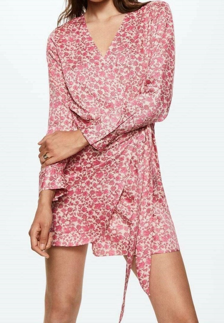 Femme portant une robe portefeuille rose à fleurs avec manches longues et col en V, nouant une ceinture en tissu à la taille, debout les bras croisés.