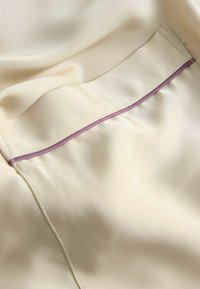 Tessuto di seta crema con una texture liscia, caratterizzato da un accento in piped viola lungo la cucitura. Include un piccolo dettaglio della tasca.