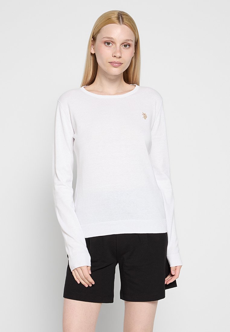 Weißer Pullover mit langen Ärmeln, Rundhalsausschnitt und geripptem Saum. Mit einem kleinen, goldenen Logo auf der Brust. Getragen mit schwarzen Shorts.