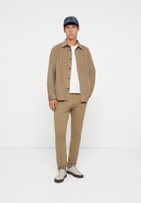 Világosbarna kockás ing, fehér póló, bézs slim-fit nadrág, szürke és fehér tornacipő, valamint kék sapka, fehér háttér előtt állva.