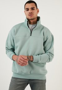 Sudadera azul claro con media cremallera, puños y cintura acanalados, textura de tela suave y un diseño en el panel frontal.