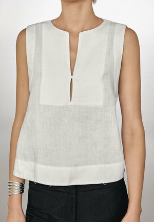 Ærmeløs hvid bluse med firkantet udskæring og frontslids, båret med sorte bukser og et sølvfarvet armbånd med flere bånd på venstre håndled.