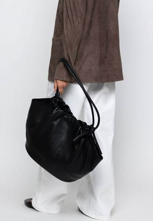 Black leather bucket bag with drawstring closure, smooth texture, and sturdy handles. Paired with a taupe suede jacket and white pants.