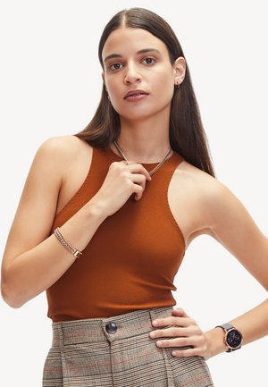 Ein rustfarbenes ärmelloses Top aus weichem Stoff, kombiniert mit karierter, hochtaillierter Hose. Die Accessoires umfassen ein silbernes Kettenarmband und eine Uhr.