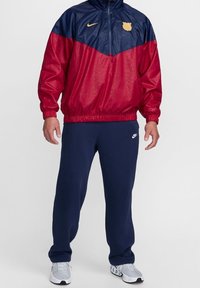 Cortavientos de Nike en rojo y azul marino, con cuello con cremallera y logo. Combinado con pantalones de chándal azul marino y zapatillas deportivas grises.
