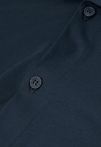 Marineblauwe stof met een gladde textuur, voorzien van twee zwarte knopen en gedetailleerde stiksel langs de rand. Eenvoudig ontwerp zonder patronen.