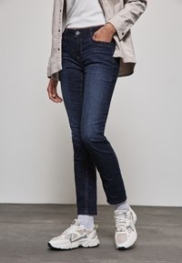 Mörk tvättade jeans med slim fit, med knappstängning och fickor; matchat med ljusgrå strumpor och kraftiga vita sneakers.