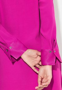 Fuchsia Bluse mit langen Ärmeln und Knopfmanschetten, die sich durch eine glatte Textur und einen Schlitz am Handgelenk auszeichnet, was das strukturierte Design des Kleidungsstücks hervorhebt.