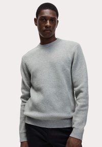 Suéter de punto gris claro con un patrón texturizado, cuello redondo y puños de canalé. La tela es suave y de peso medio.
