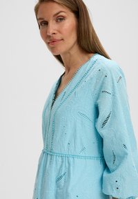 Helles blaues besticktes Kleid mit V-Ausschnitt, langen Ärmeln und dekorativer Naht. Es verfügt über komplexe Muster und einen elastischen Taillenbund.