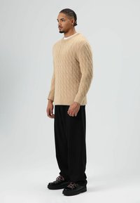 Beige kabelgebreide trui met geribbelde manchetten, gecombineerd met losse zwarte broek en chunky zwarte schoenen met roze veters.