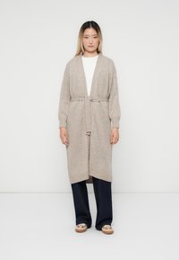 Beige gestrickte Strickjacke mit einem strukturierten Muster, offener Front und einem Bindegurt in der Taille. Über einem weißen Oberteil getragen und mit dunklen Hosen kombiniert.