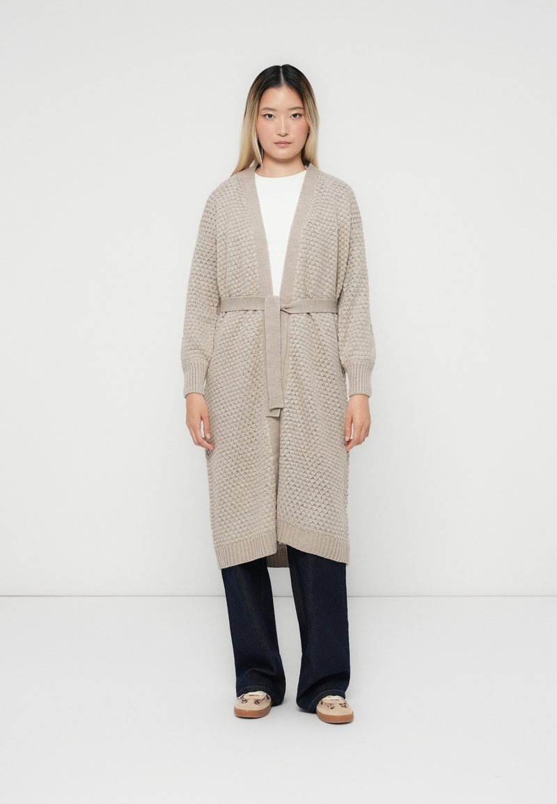 Beige gestrickte Strickjacke mit einem strukturierten Muster, offener Front und einem Bindegurt in der Taille. Über einem weißen Oberteil getragen und mit dunklen Hosen kombiniert.