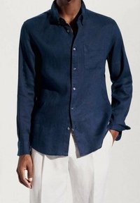 Chemise bleu marine à boutons, en tissu texturé, dotée d'une poche plaquée, de manches longues et de boutons blancs. Associée à un pantalon de couleur claire.