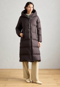 Femme portant un long manteau rembourré marron foncé avec un col haut, un pantalon beige évasé et des bottines à talons crèmes, se tenant sur un sol en bois.