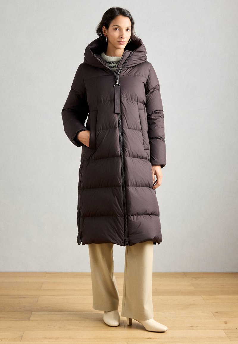 Femme portant un long manteau rembourré marron foncé avec un col haut, un pantalon beige évasé et des bottines à talons crèmes, se tenant sur un sol en bois.